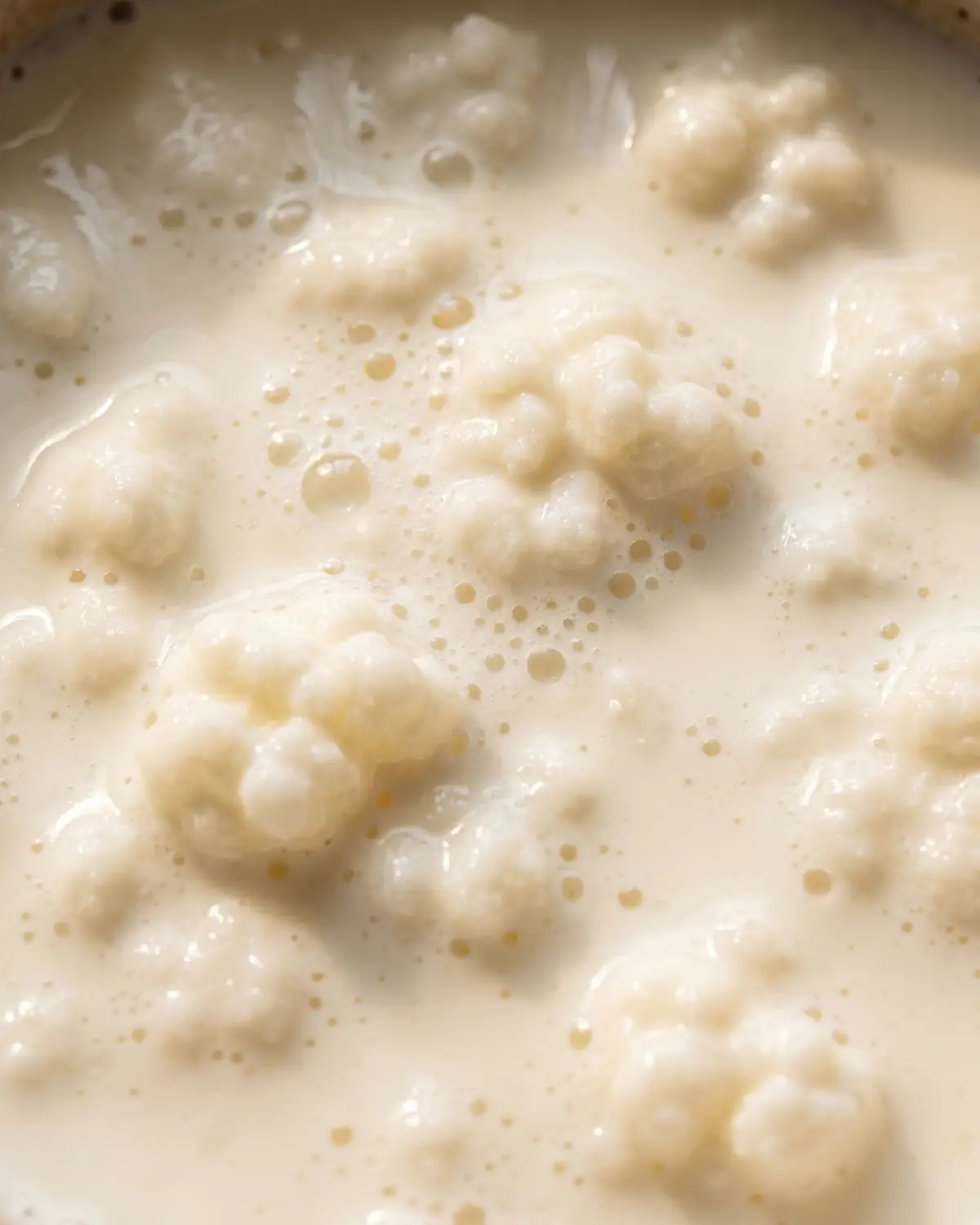 Superficie cremosa del k&eacute;fir de leche vista de cerca, con burbujas finas bajo una luz cenital suave