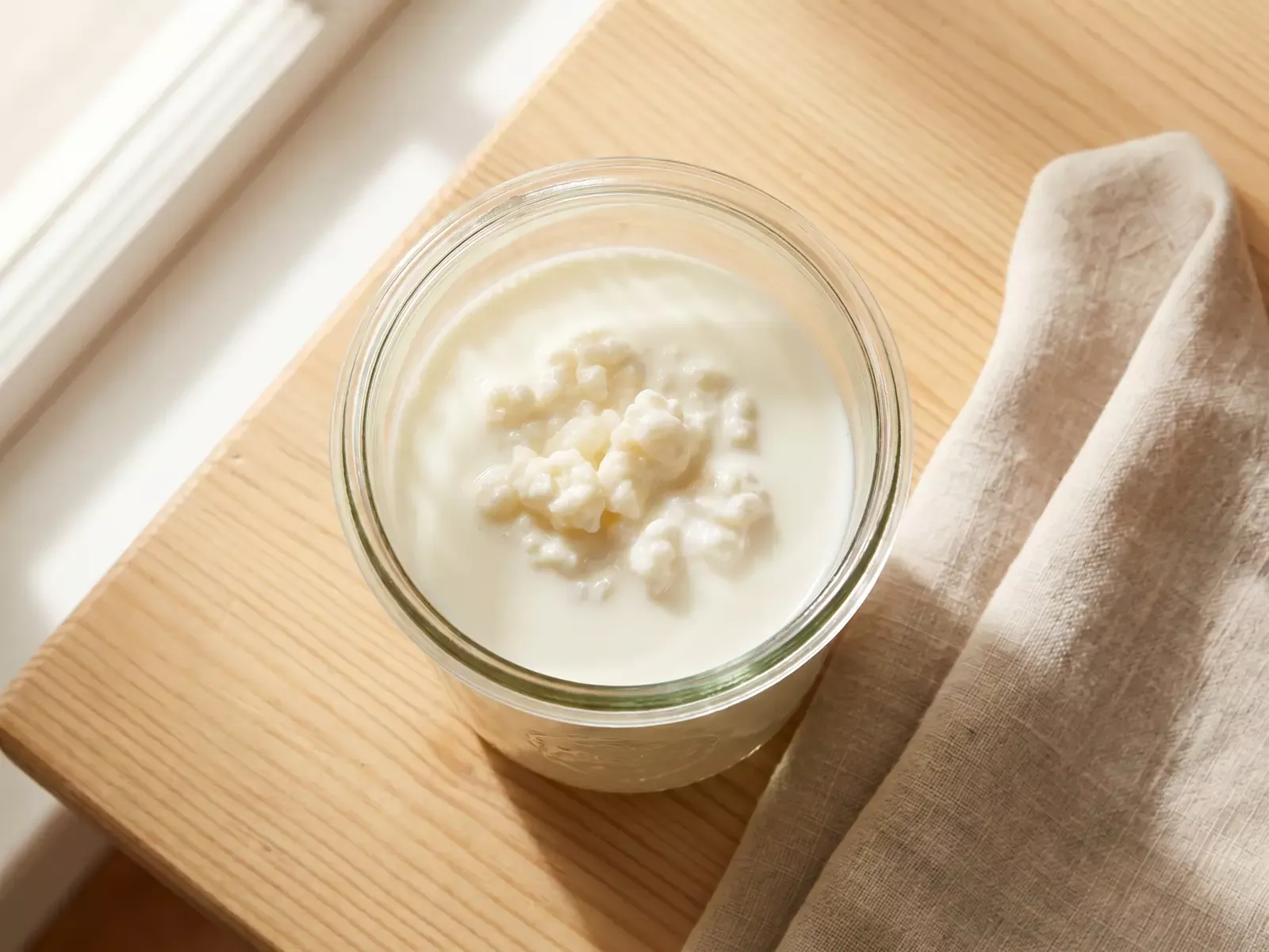 alimentos fermentados Tarro de cristal con kéfir de leche fermentando sobre madera, junto a un paño de lino en vista cenital.