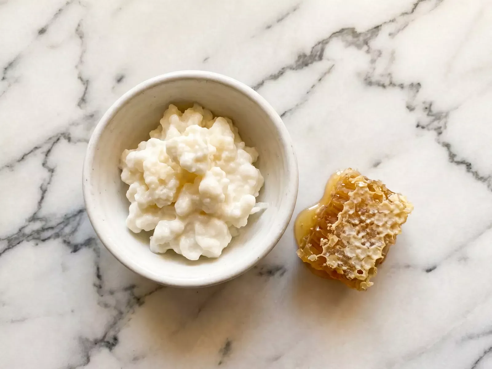 leche fermentada Cuenco de kéfir natural con panal de miel sobre mármol veteado y luz cenital cálida.