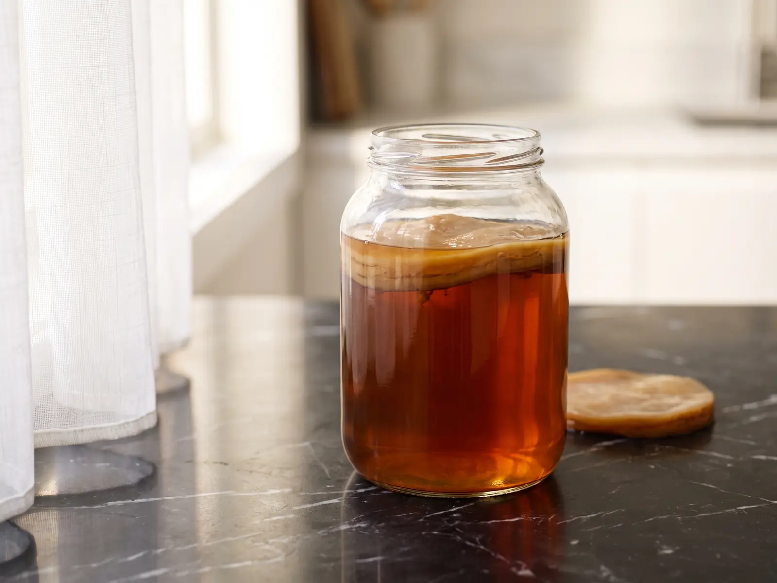 kombucha opiniones Tarro con SCOBY sobre mármol negro y té ámbar profundo iluminado por una cortina blanca.