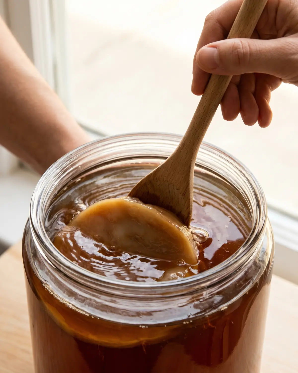 Cuchara de madera empujando suavemente un SCOBY bajo t&eacute; &aacute;mbar durante la preparaci&oacute;n de kombucha casera
