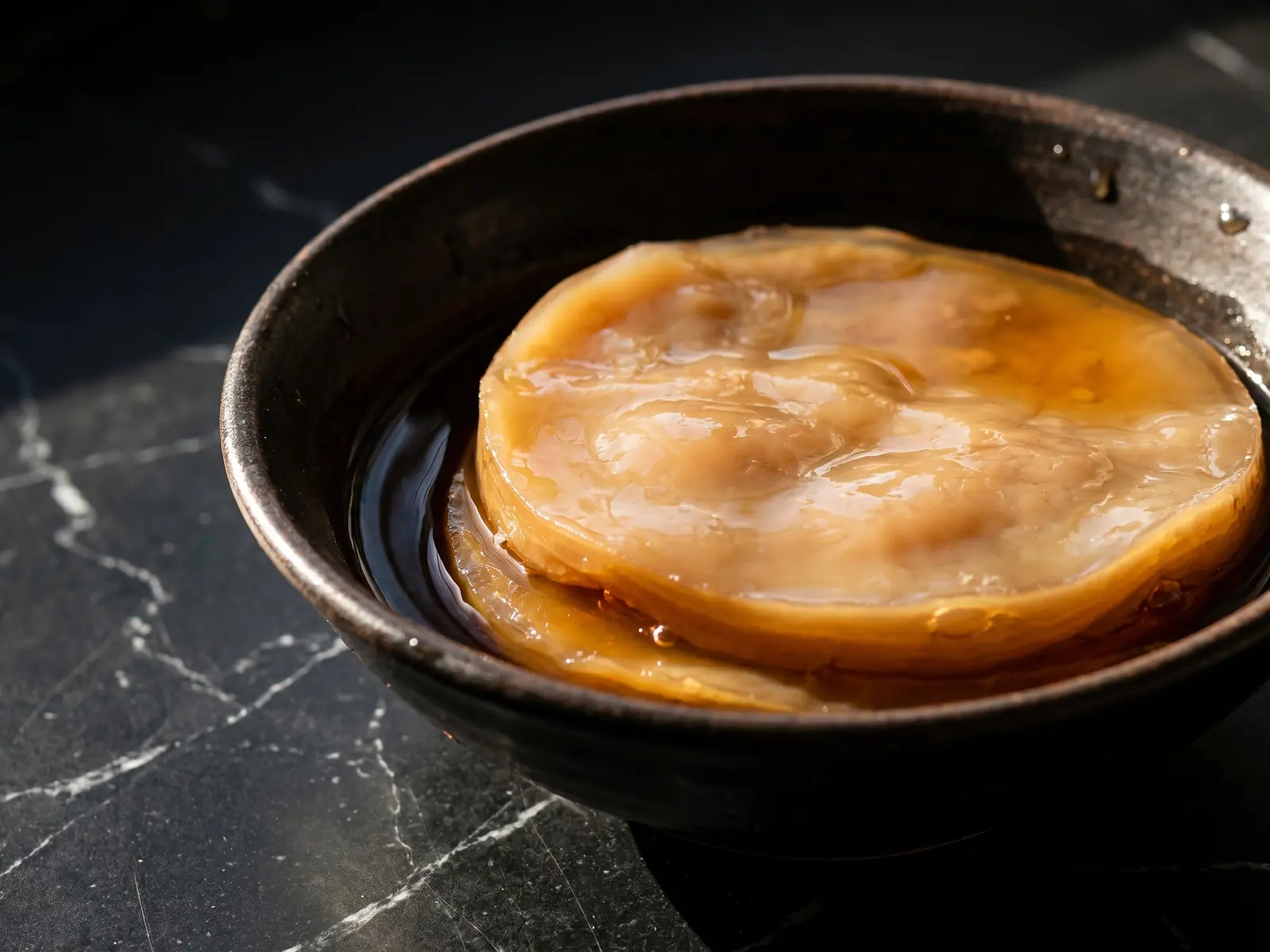 Cuenco de t&eacute; oscuro con un SCOBY beige sobre m&aacute;rmol negro mate iluminado con luz natural