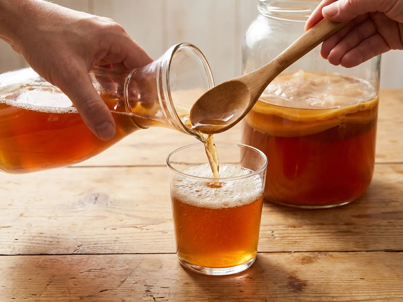 como hacer kombucha Mano sirviendo kombucha terminada en un vaso de cristal con espuma fina sobre una mesa de pino.