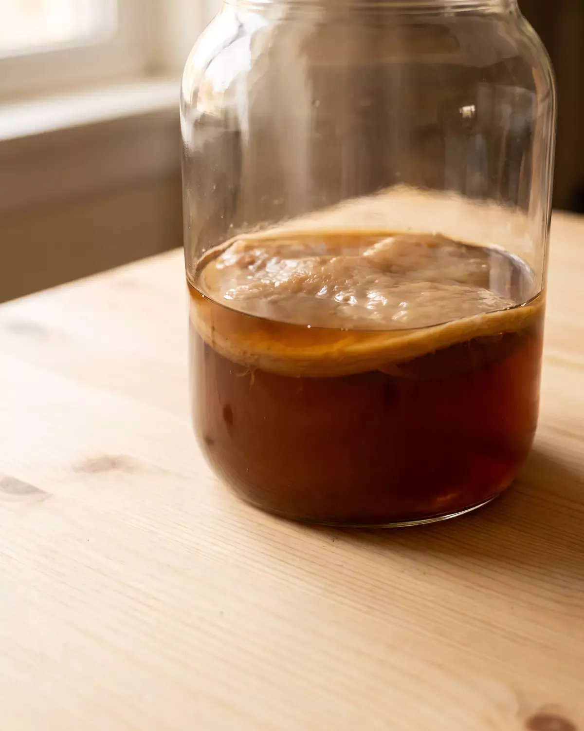 Vista vertical de un tarro de kombucha donde el SCOBY &aacute;mbar destaca sobre una base de pino claro