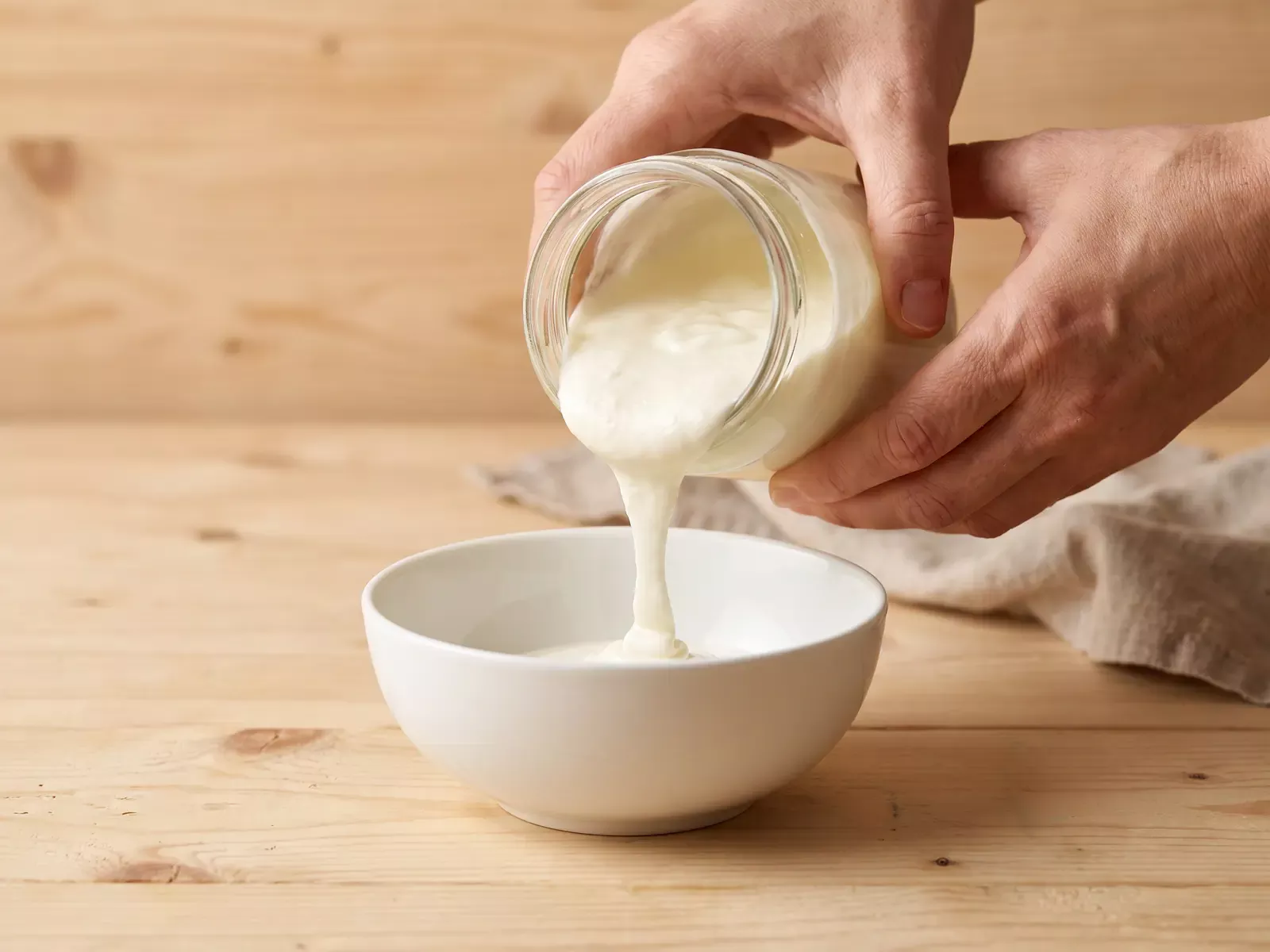 Dos manos vierten un yogur espeso desde un tarro hacia un cuenco blanco sobre una mesa de pino claro.